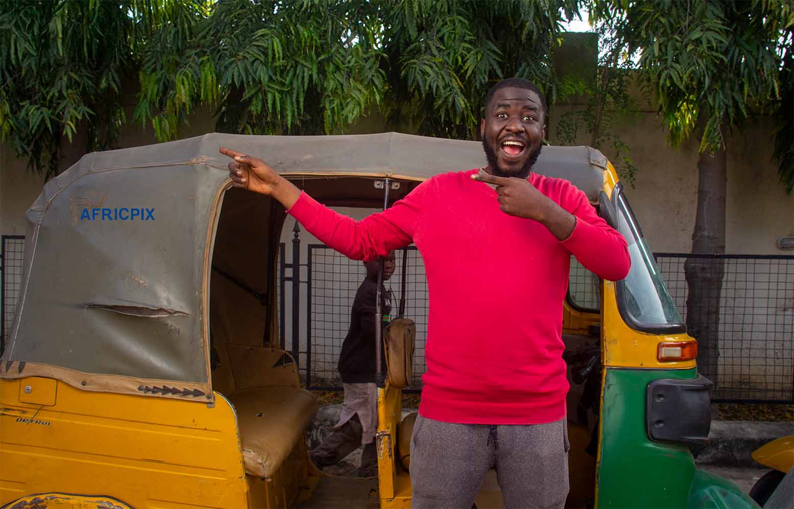 A happy African tricycle,Keke, Maruwa rider  standing with his tricycle behind him, pointing to the left with a cheerful expression.